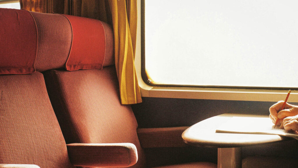 Train seat, train window and a hand holding a pen in the act of writing 